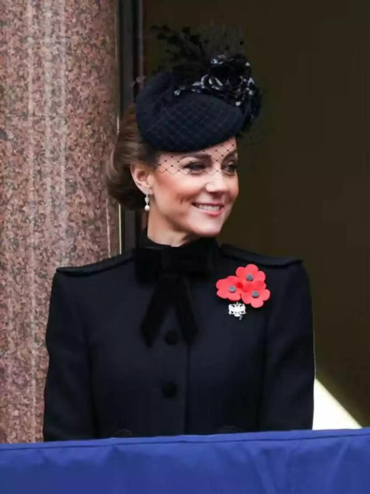 Kate Middleton sonriente con un sombrero negro elegante y un abrigo oscuro adornado con amapolas rojas.