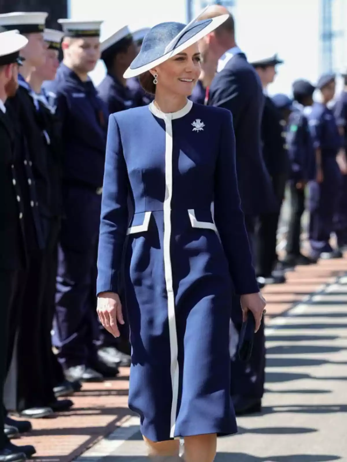 Una mujer con un elegante vestido azul y sombrero a juego camina al aire libre mientras personas uniformadas están alineadas a su alrededor. Una mujer con un elegante vestido azul y sombrero a juego camina al aire libre mientras personas uniformadas están alineadas a su alrededor.