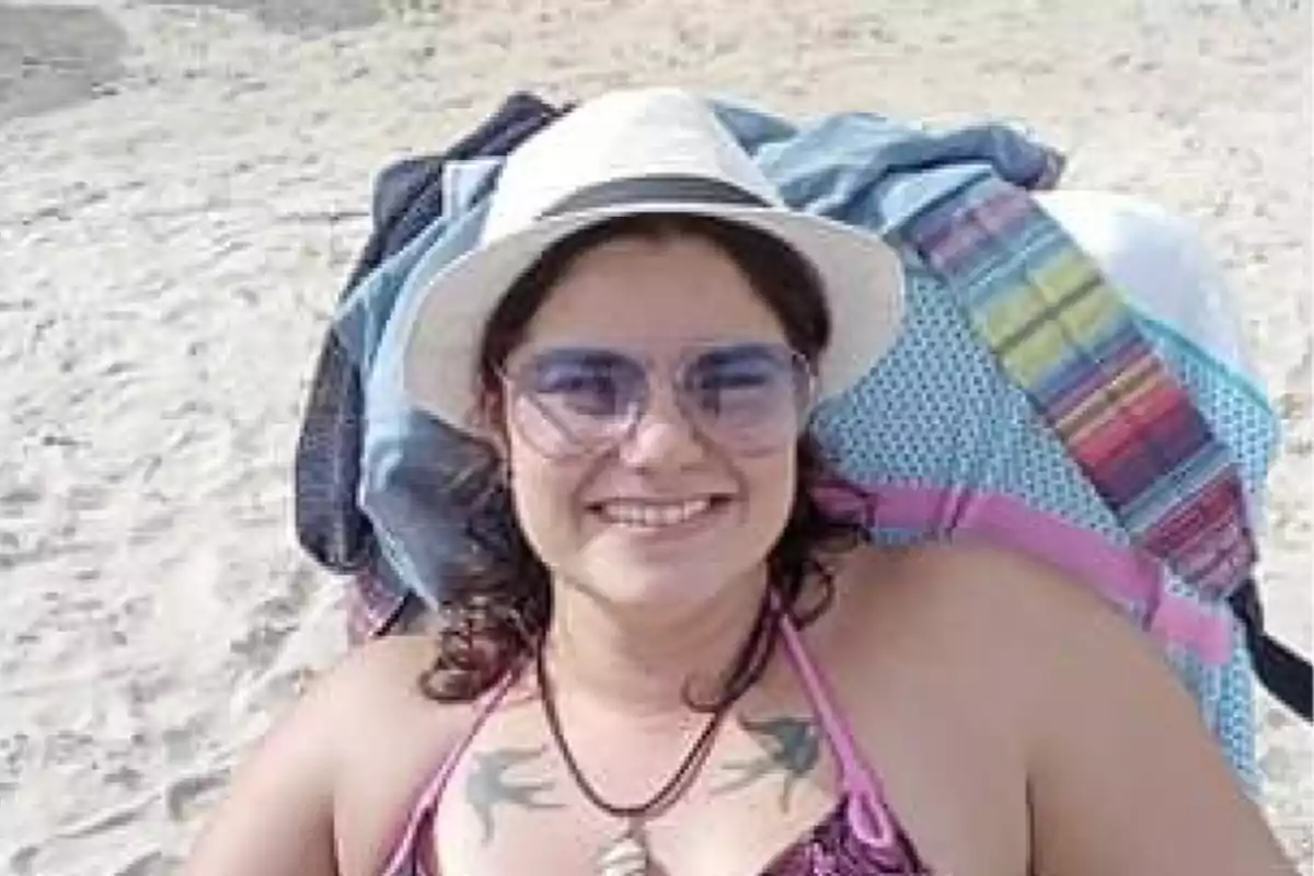 Mujer sonriente con sombrero blanco y gafas de sol, recostada en una silla de playa sobre la arena.