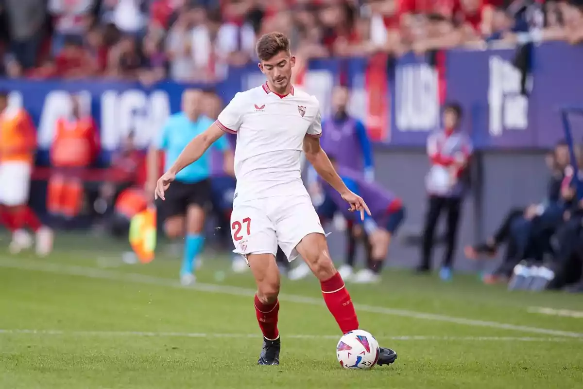 Jugador de fútbol del Sevilla FC controlando el balón en un partido con espectadores y árbitros de fondo.