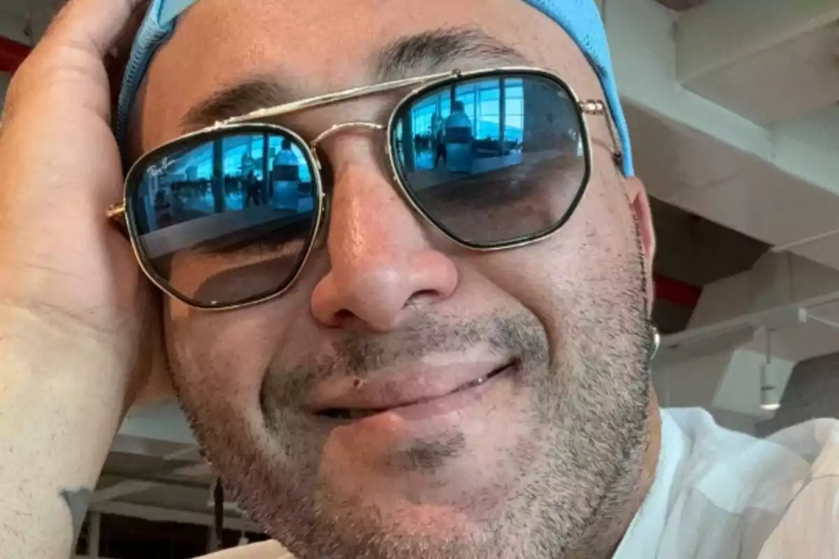 Un hombre con gorra azul y gafas de sol sonríe mientras apoya su cabeza en su mano. Un hombre con gorra azul y gafas de sol sonríe mientras apoya su cabeza en su mano.
