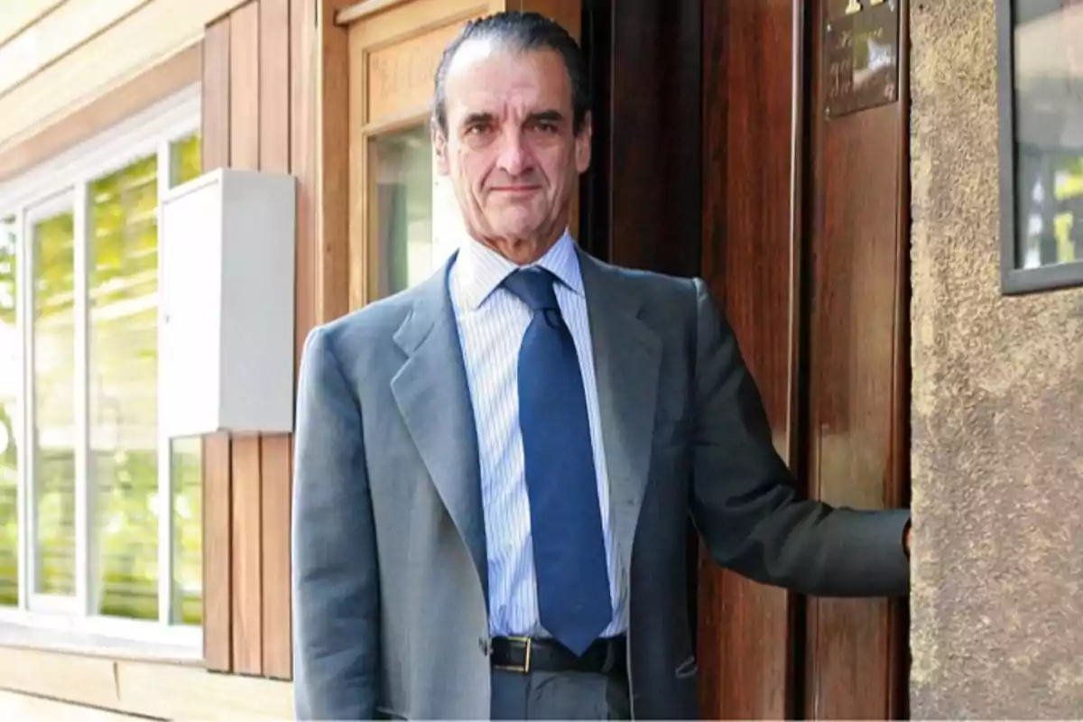 Un hombre de traje gris y corbata azul está de pie junto a una puerta de madera.