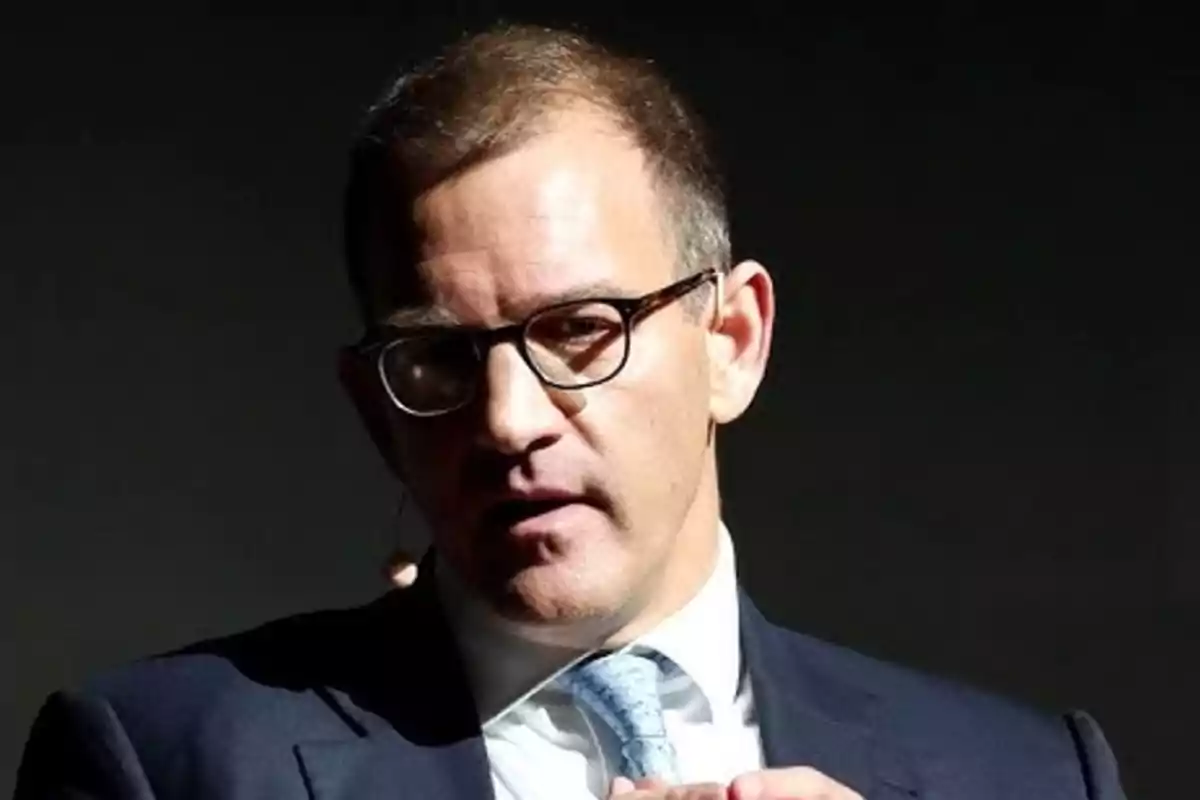 Un hombre con gafas y traje oscuro hablando en un escenario iluminado. Un hombre con gafas y traje oscuro hablando en un escenario iluminado.