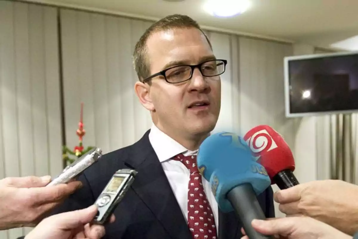 Un hombre con gafas y traje oscuro es entrevistado por varios periodistas que sostienen micrófonos de diferentes colores. Un hombre con gafas y traje oscuro es entrevistado por varios periodistas que sostienen micrófonos de diferentes colores.