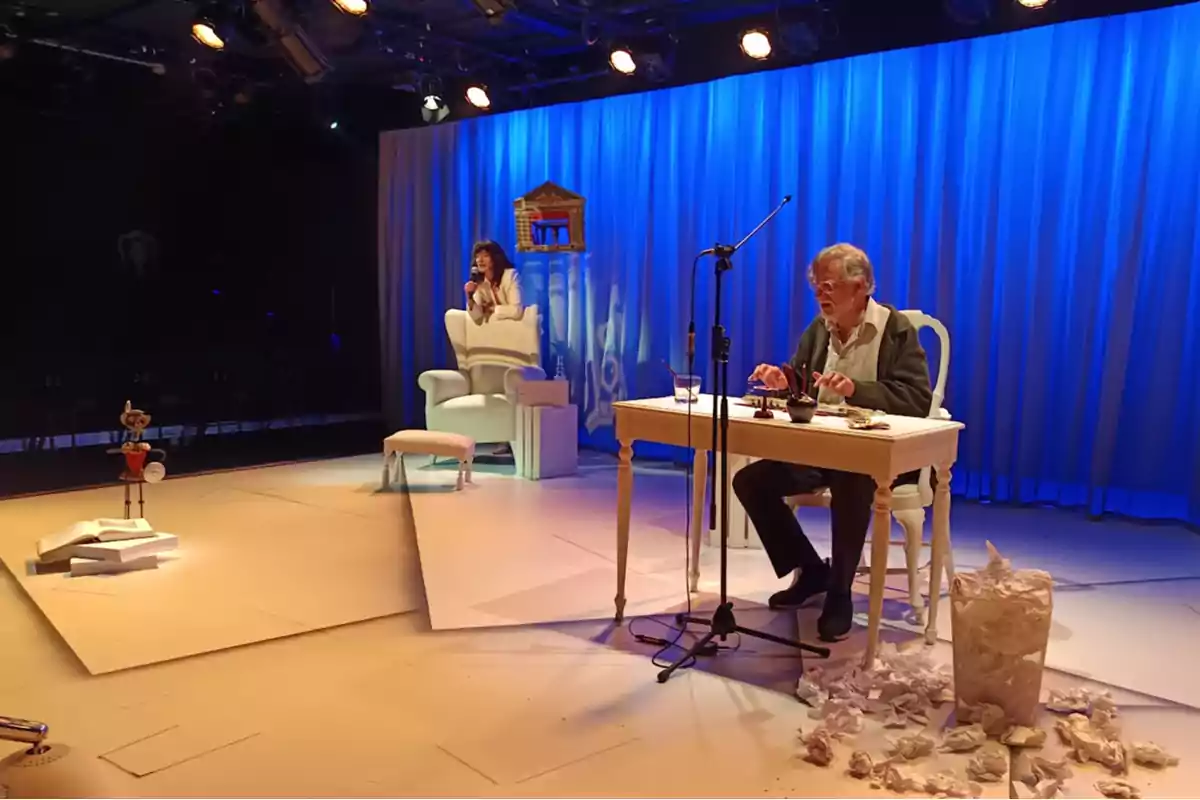 Un hombre mayor está sentado escribiendo en una mesa con papeles arrugados en el suelo mientras una mujer está sentada en un sillón al fondo y una marioneta de Pinocho está de pie sobre un libro gigante en un escenario iluminado con luz azul Un hombre mayor está sentado escribiendo en una mesa con papeles arrugados en el suelo mientras una mujer está sentada en un sillón al fondo y una marioneta de Pinocho está de pie sobre un libro gigante en un escenario iluminado con luz azul