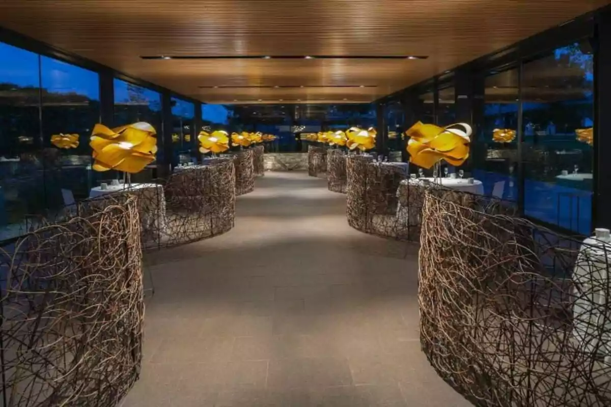 Un elegante restaurante con mesas separadas por estructuras de ramas entrelazadas y lámparas decorativas amarillas.