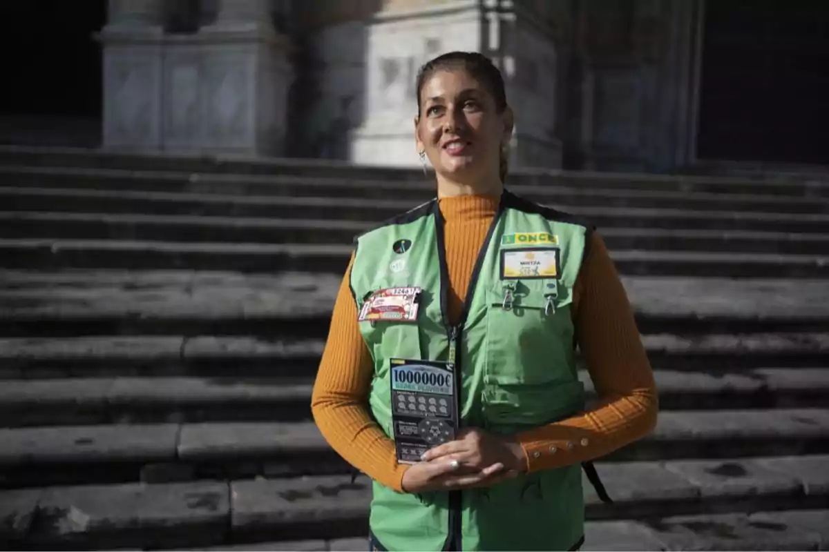 Una persona con chaleco verde de la ONCE sostiene un cupón de lotería frente a unas escaleras.