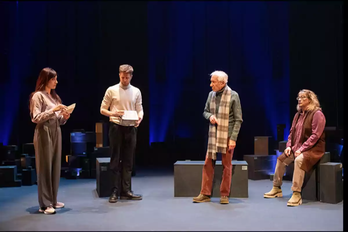 Cuatro personas en un escenario teatral, dos de pie y dos sentadas, con cajas alrededor y un fondo oscuro iluminado.