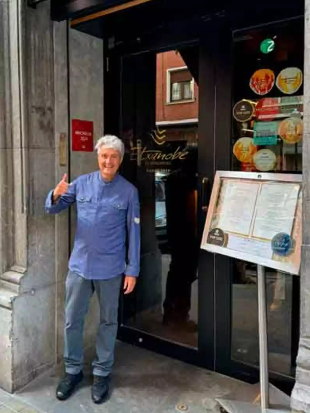 Una persona sonriente con una chaqueta azul está de pie frente a la entrada de un restaurante llamado 