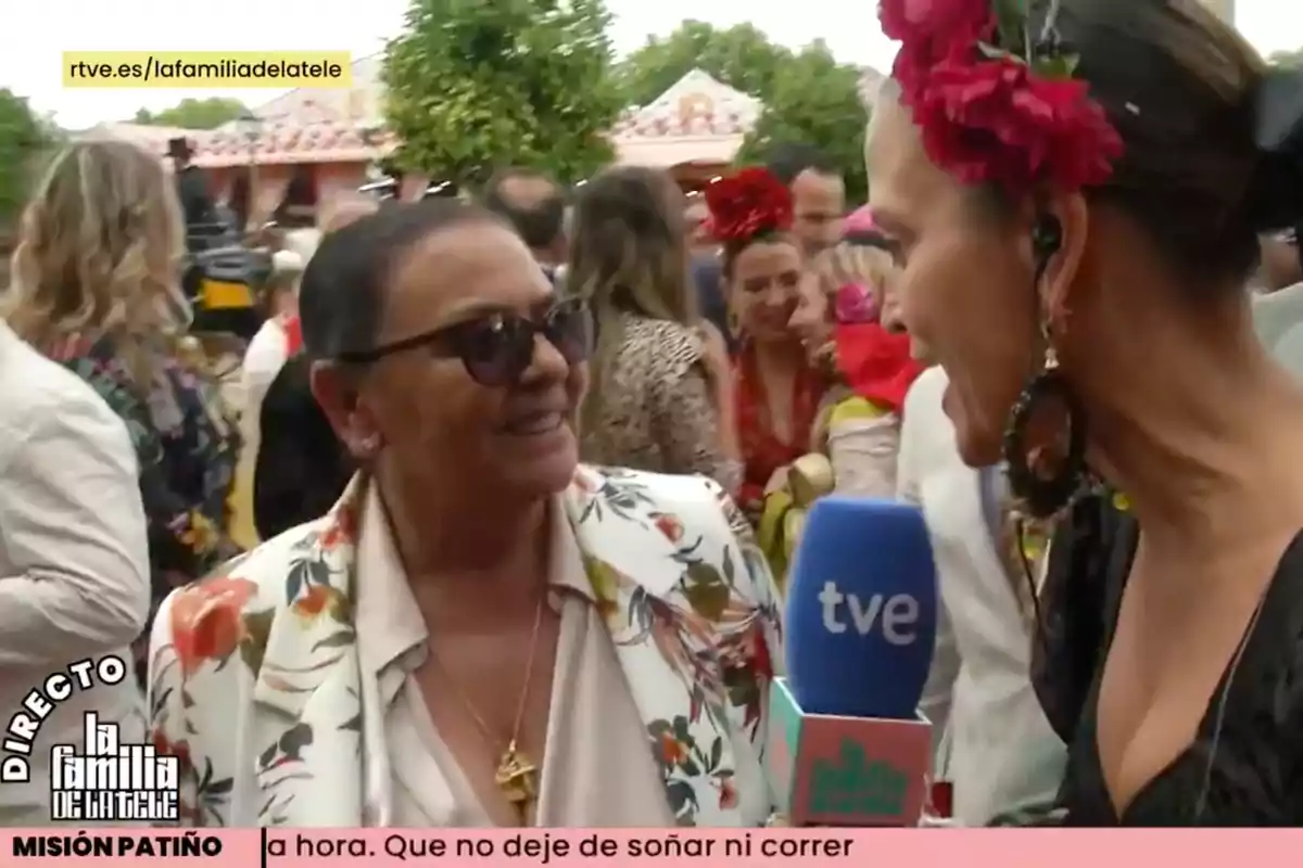 Una reportera entrevista a María del Monte en un evento al aire libre, rodeadas de personas vestidas con trajes coloridos y adornos florales. Captura de 'La Familia de la Tele' Una reportera entrevista a María del Monte en un evento al aire libre, rodeadas de personas vestidas con trajes coloridos y adornos florales. Captura de 'La Familia de la Tele'