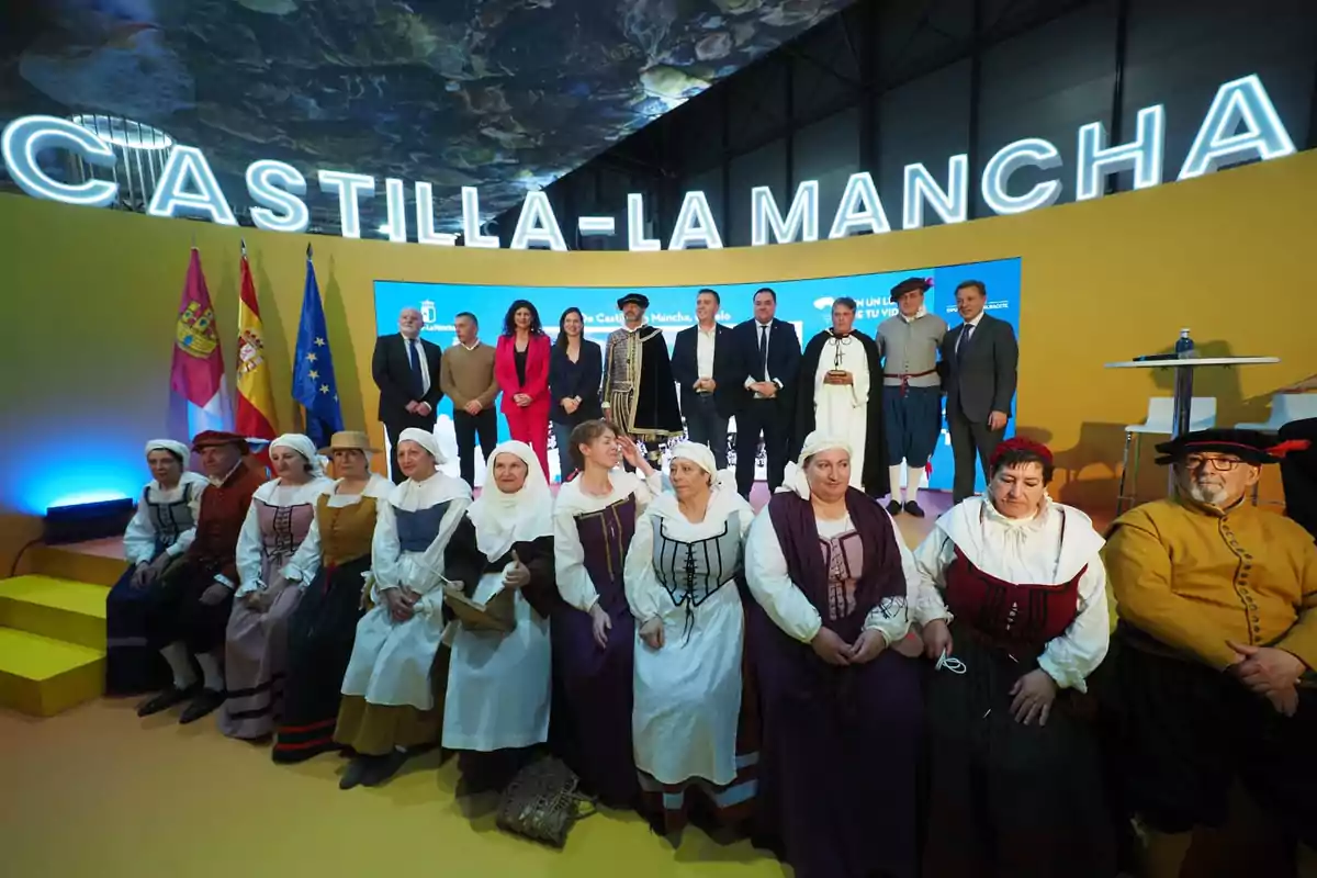 Un grupo de personas posa en un escenario con el letrero de Castilla-La Mancha, algunas vestidas con trajes tradicionales, frente a banderas y un fondo iluminado.