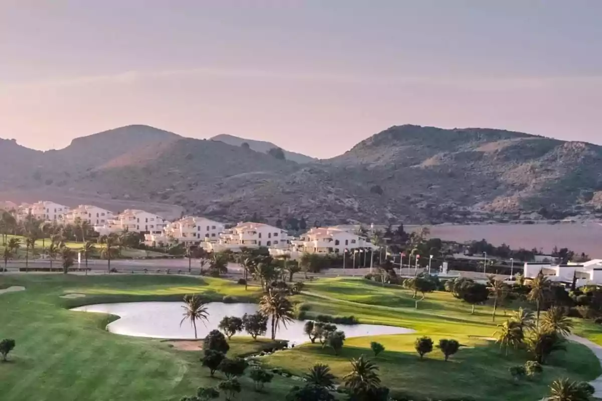 Vista de un campo de golf con un lago pequeño, rodeado de palmeras y casas blancas al fondo, con montañas en el horizonte bajo un cielo despejado.