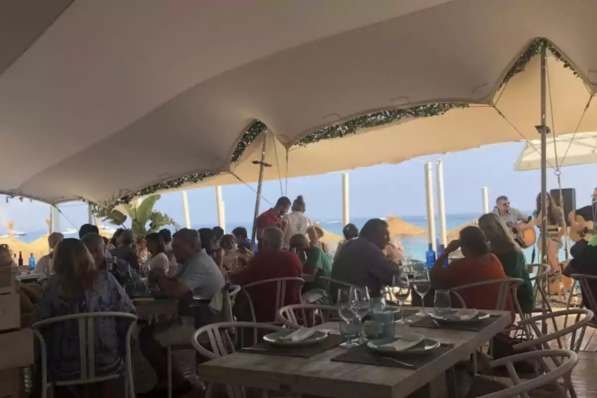 Personas disfrutando de una comida en un restaurante al aire libre con vista al mar y música en vivo.