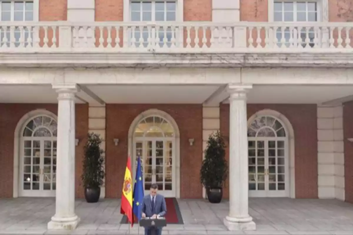 Un hombre de traje da un discurso frente a un atril entre las banderas de España y la Unión Europea en la entrada de un edificio oficial con columnas y grandes ventanales. Un hombre de traje da un discurso frente a un atril entre las banderas de España y la Unión Europea en la entrada de un edificio oficial con columnas y grandes ventanales.