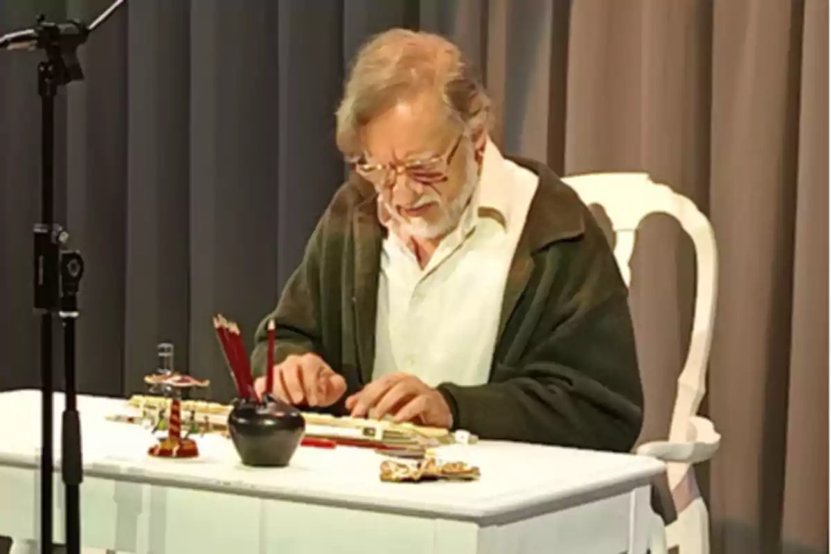 Hombre mayor con gafas sentado en una mesa blanca escribiendo con pluma rodeado de objetos antiguos Hombre mayor con gafas sentado en una mesa blanca escribiendo con pluma rodeado de objetos antiguos