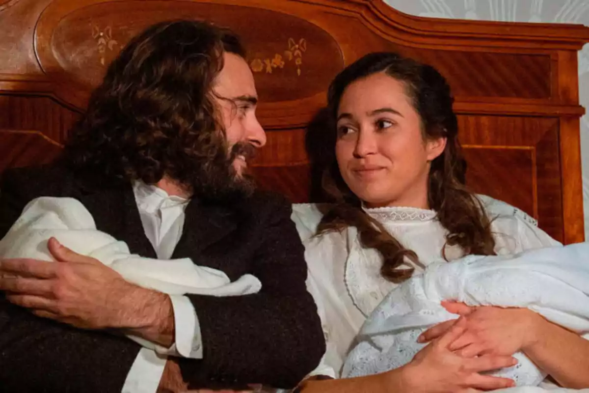Adriano y Catalina sonriente en la cama, cada uno sosteniendo a un bebé envuelto en mantas blancas. Captura de 'la Promesa' Adriano y Catalina sonriente en la cama, cada uno sosteniendo a un bebé envuelto en mantas blancas. Captura de 'la Promesa'