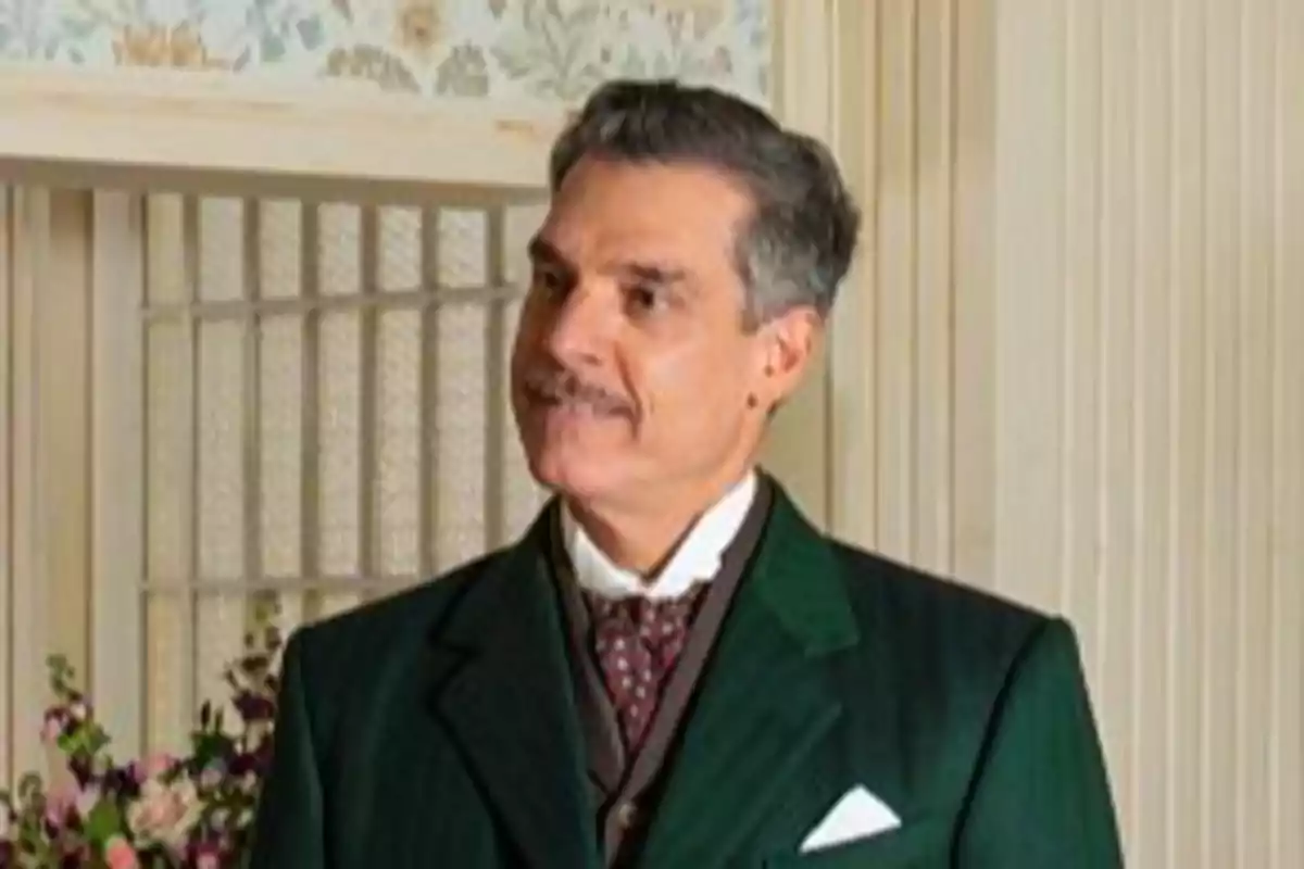 Lisandro con bigote y cabello canoso lleva un traje verde oscuro y corbata, de pie frente a una pared decorada con papel tapiz floral. Captura de 'La Promesa' Lisandro con bigote y cabello canoso lleva un traje verde oscuro y corbata, de pie frente a una pared decorada con papel tapiz floral. Captura de 'La Promesa'