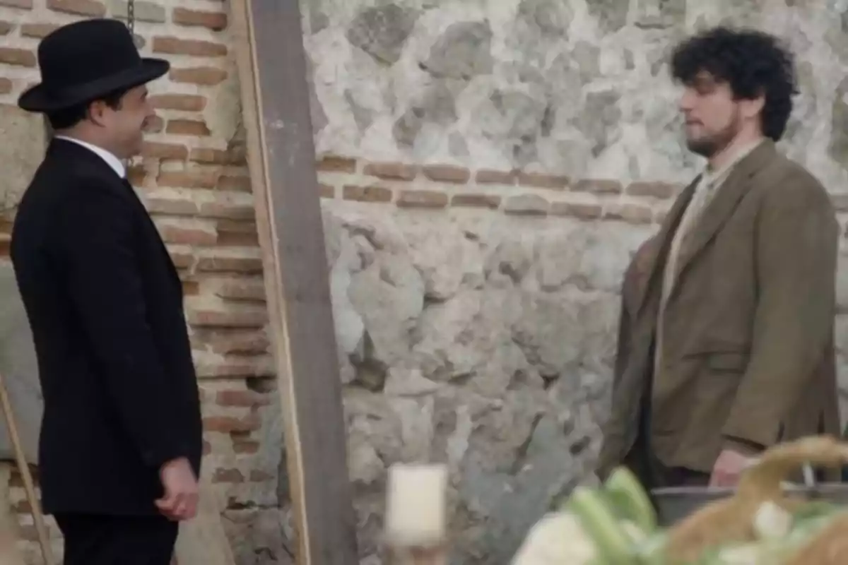 Manuel y Toño conversan frente a una pared de piedra, uno lleva un traje negro y sombrero, el otro viste un traje marrón. Captura de 'La Promesa' Manuel y Toño conversan frente a una pared de piedra, uno lleva un traje negro y sombrero, el otro viste un traje marrón. Captura de 'La Promesa'