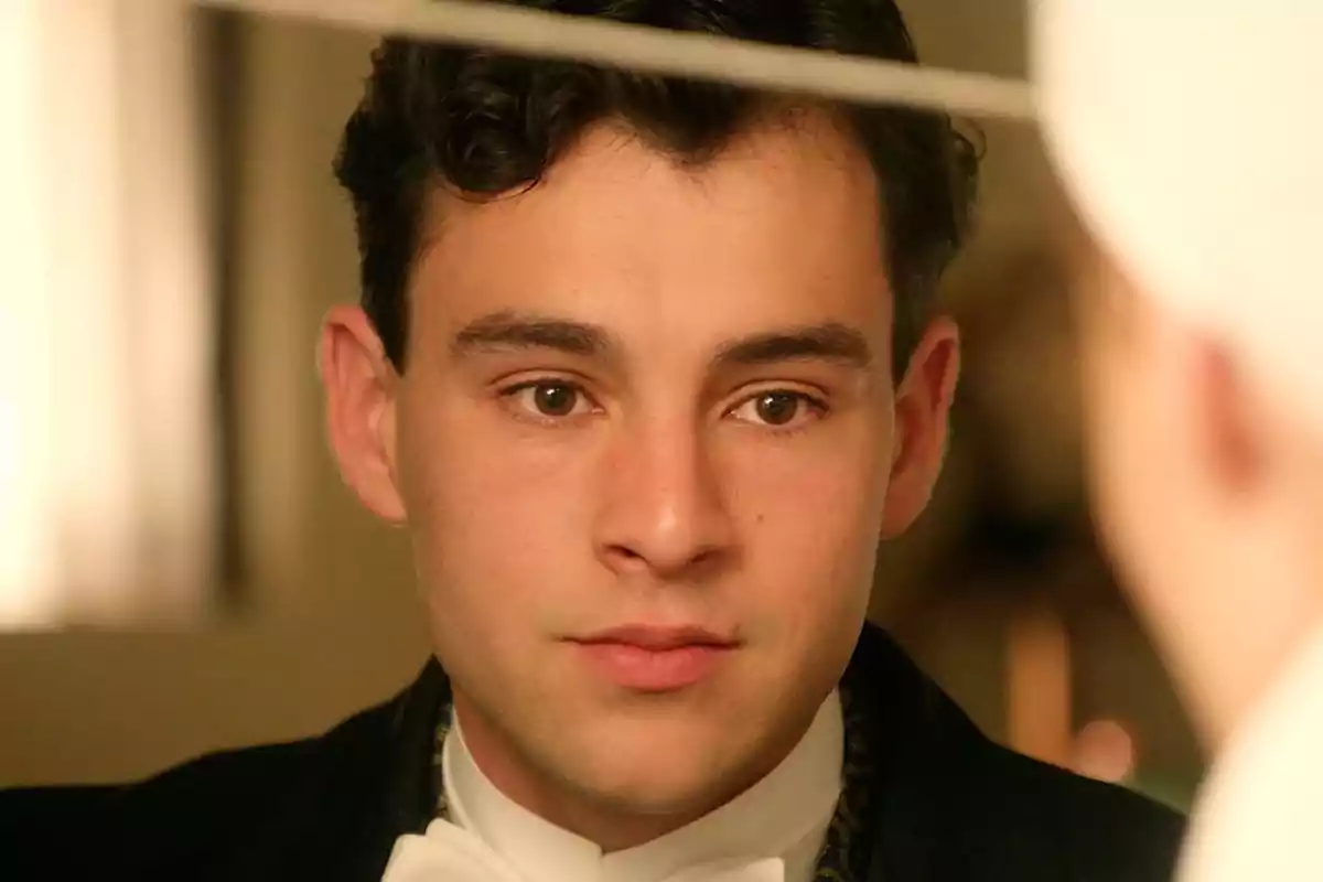 Curro con cabello oscuro y rizado viste un traje formal y mira hacia adelante con expresión seria. Captura de 'La Promesa' Curro con cabello oscuro y rizado viste un traje formal y mira hacia adelante con expresión seria. Captura de 'La Promesa'