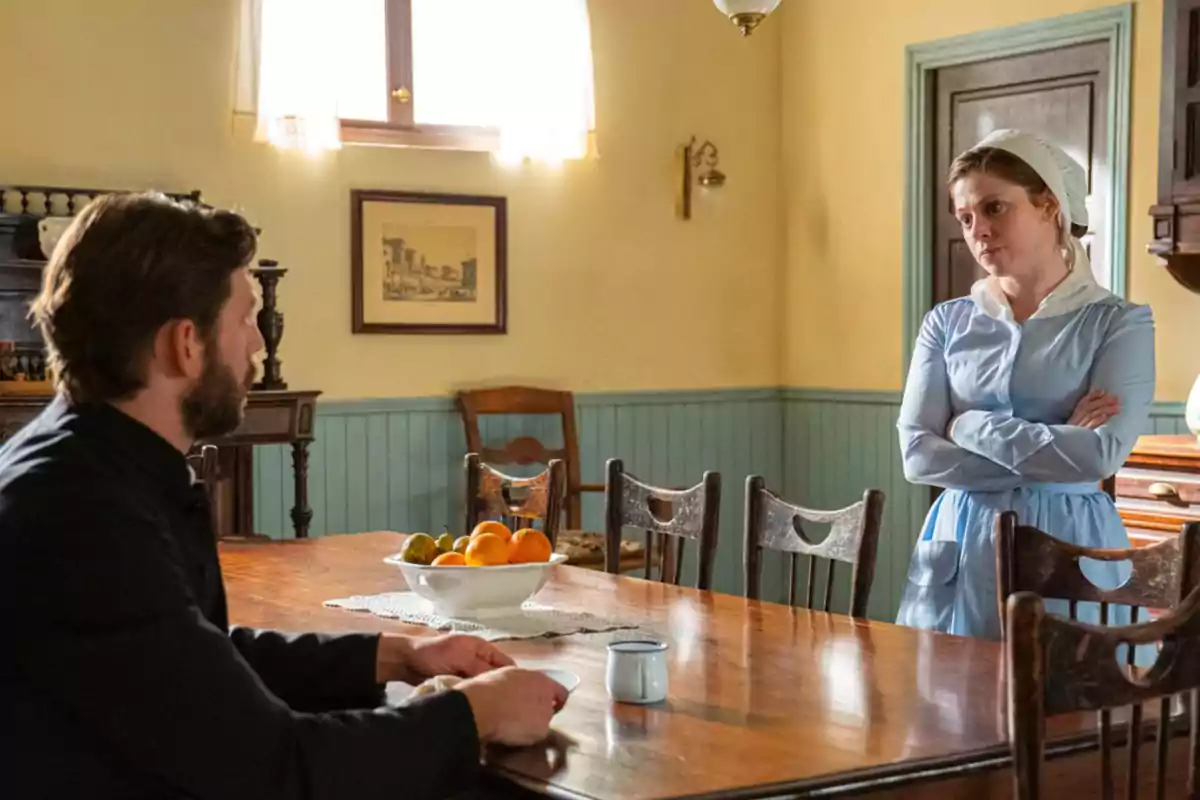 Samuel sentado a la mesa de un comedor antiguo conversa con María vestida de sirvienta que está de pie con los brazos cruzados. Captura de 'La Promesa' Samuel sentado a la mesa de un comedor antiguo conversa con María vestida de sirvienta que está de pie con los brazos cruzados. Captura de 'La Promesa'