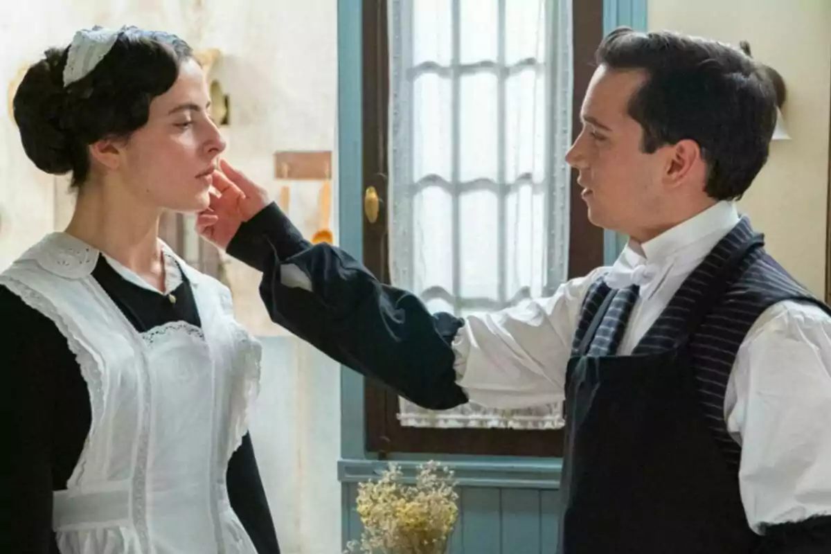Vera y Santos vestidos con ropa de época, Vera con uniforme de sirvienta y Santos con chaleco y pajarita, interactúan en una habitación iluminada. Captura de 'La Promesa' Vera y Santos vestidos con ropa de época, Vera con uniforme de sirvienta y Santos con chaleco y pajarita, interactúan en una habitación iluminada. Captura de 'La Promesa'