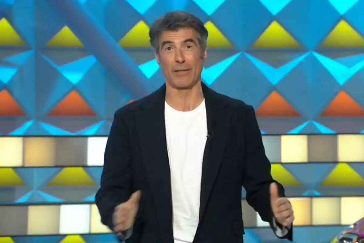 Jorge Fernández con chaqueta negra y camiseta blanca está de pie frente a un fondo colorido con patrones geométricos. Captura de 'La Ruleta de la Suerte' Jorge Fernández con chaqueta negra y camiseta blanca está de pie frente a un fondo colorido con patrones geométricos. Captura de 'La Ruleta de la Suerte'