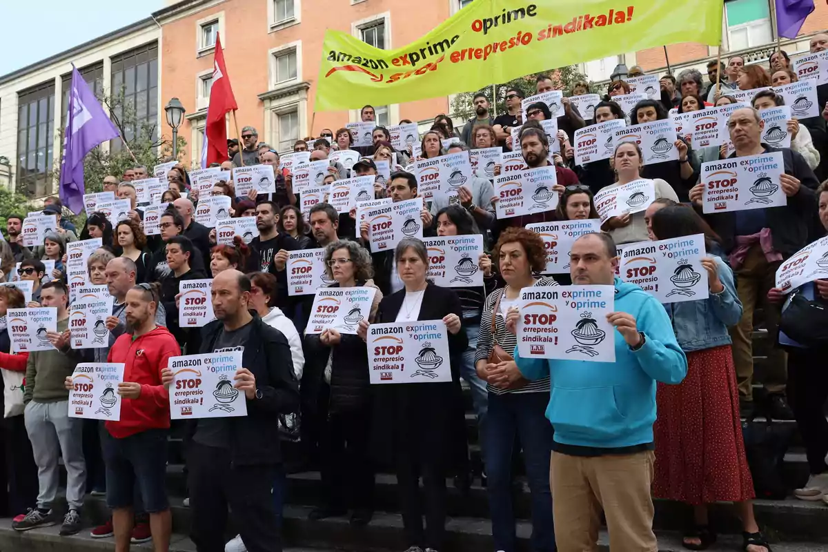 Un grupo de personas se manifiesta sosteniendo carteles que critican a una empresa, con banderas y pancartas en el fondo.