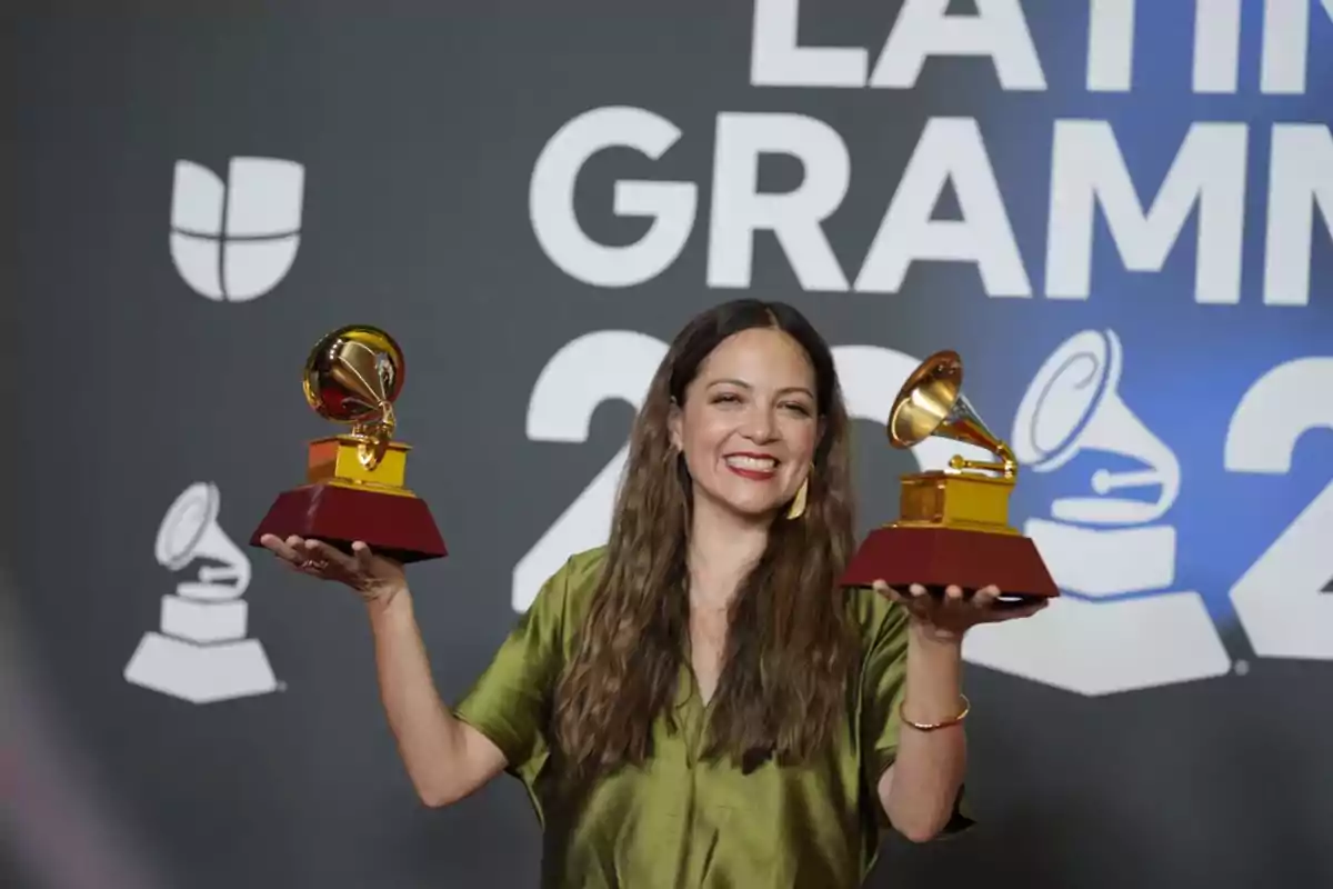 Una persona sonriente sostiene dos premios Grammy Latinos frente a un fondo con el logo del evento.