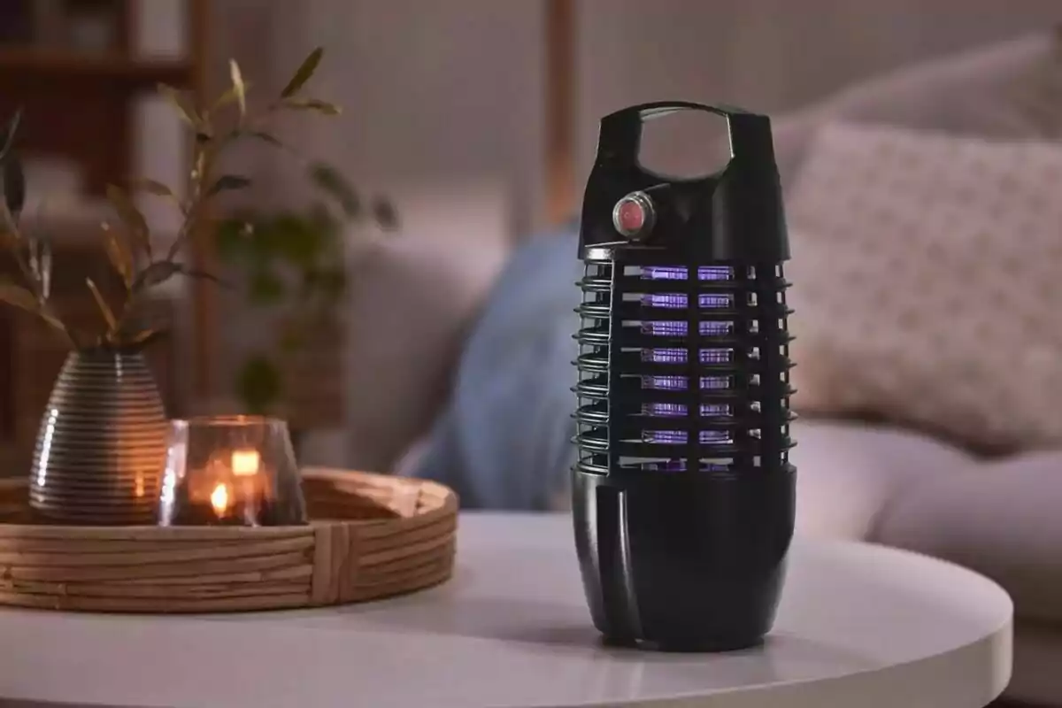 Un dispositivo negro con luz violeta en una mesa blanca junto a una bandeja de mimbre con una vela encendida y un jarrón con ramas. Un dispositivo negro con luz violeta en una mesa blanca junto a una bandeja de mimbre con una vela encendida y un jarrón con ramas.