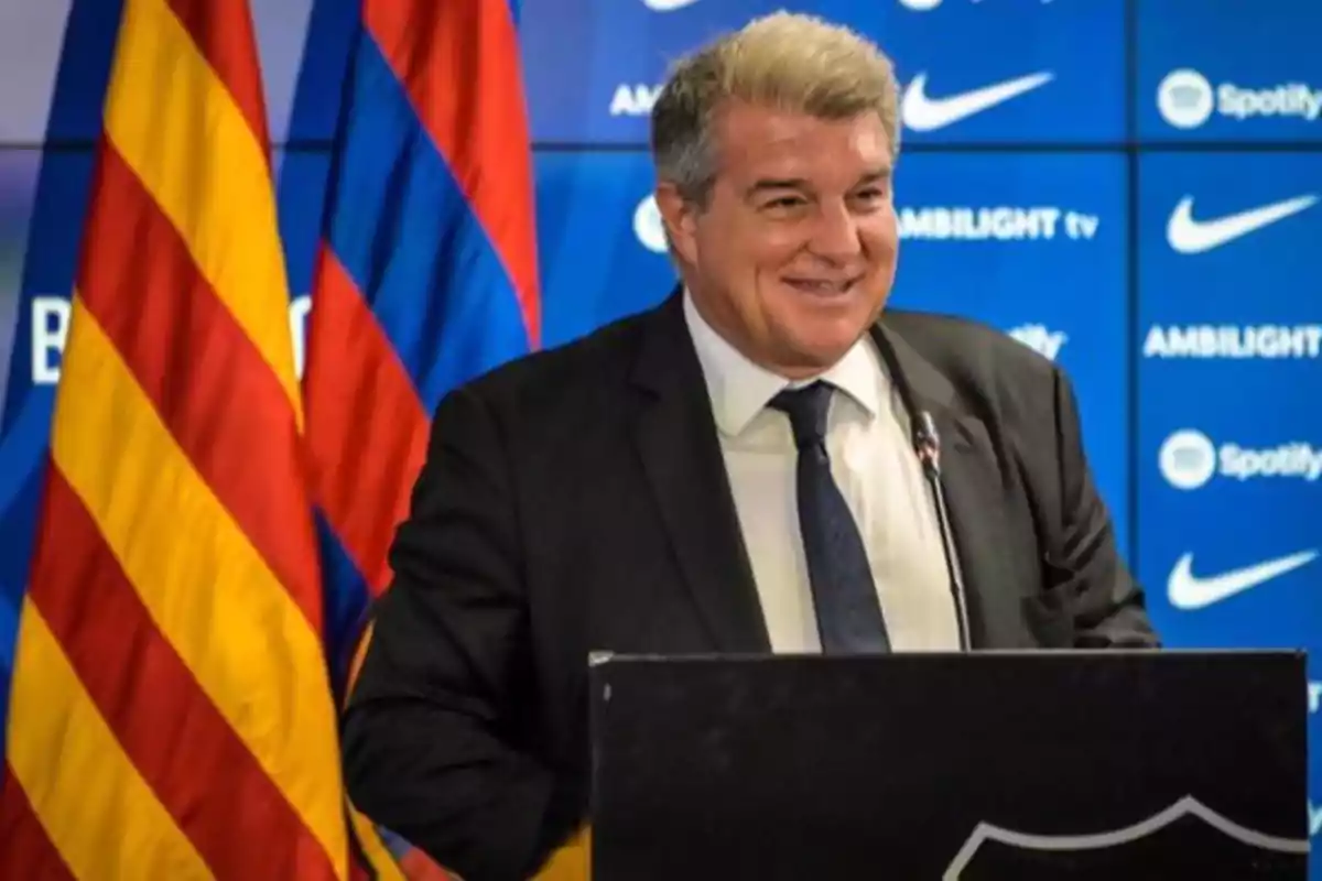 Hombre sonriendo en una conferencia de prensa con banderas y logotipos de fondo.