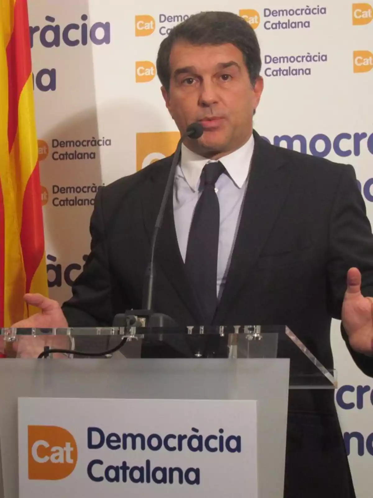 Hombre en traje hablando en un podio con el logotipo de Democràcia Catalana y una bandera catalana al fondo.