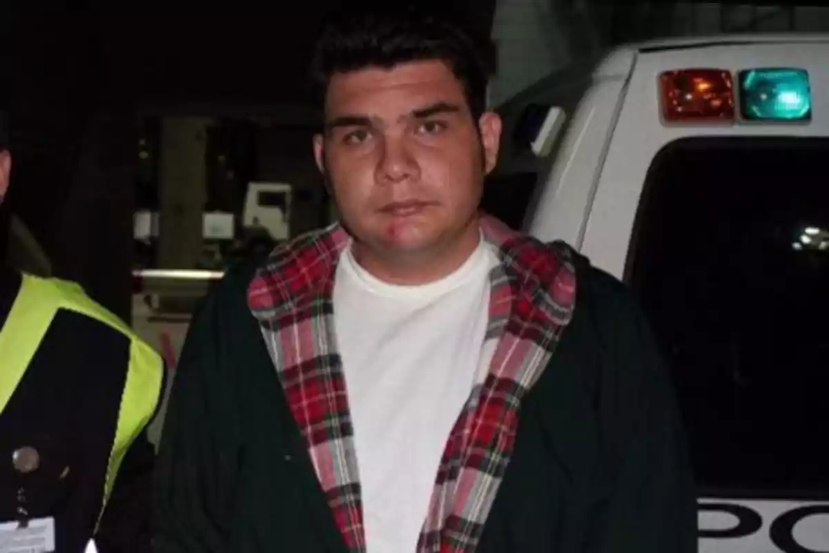 Un hombre con chaqueta de cuadros y camiseta blanca está de pie frente a un vehículo policial.