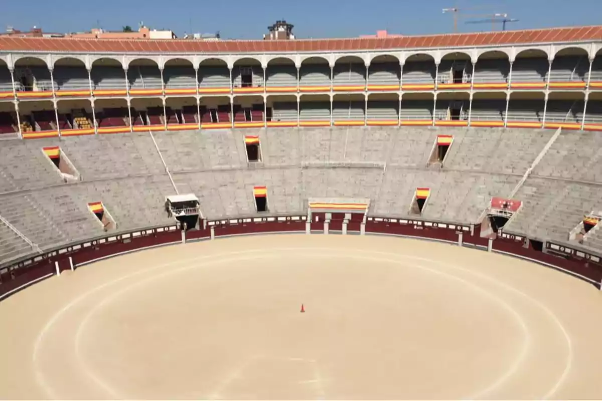 Una plaza de toros vacía con gradas y arcos, decorada con banderas.