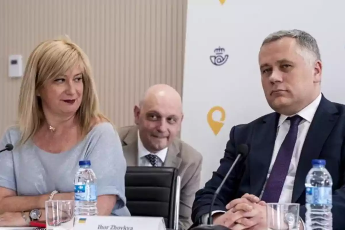 Tres personas sentadas en una mesa de conferencia, dos de ellas al frente con expresión seria y una en el centro al fondo sonriendo, con botellas de agua y micrófonos sobre la mesa Tres personas sentadas en una mesa de conferencia, dos de ellas al frente con expresión seria y una en el centro al fondo sonriendo, con botellas de agua y micrófonos sobre la mesa