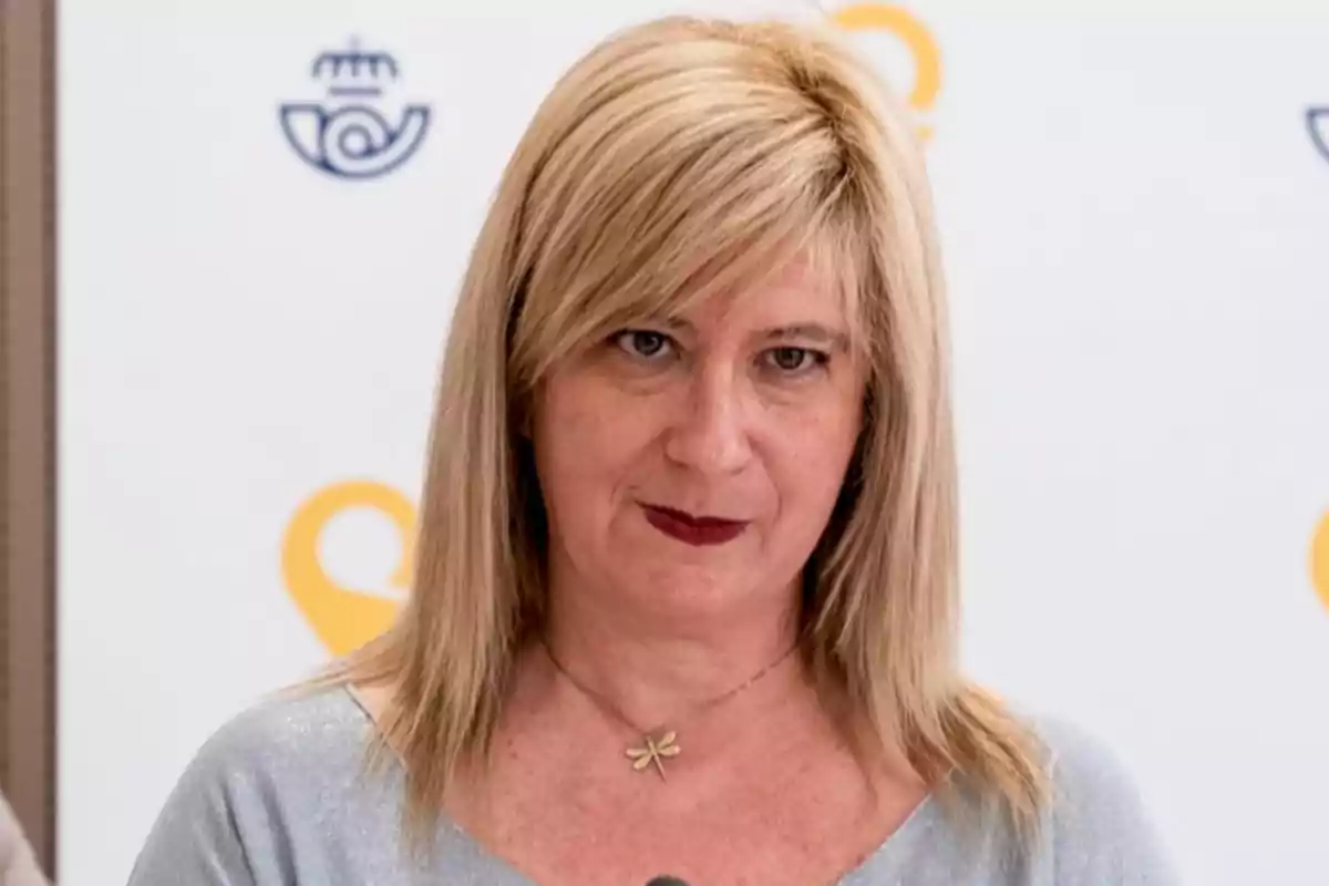 Mujer rubia de cabello lacio y suelto con labios pintados de rojo y collar de libélula frente a un fondo blanco con logotipos azules y amarillos