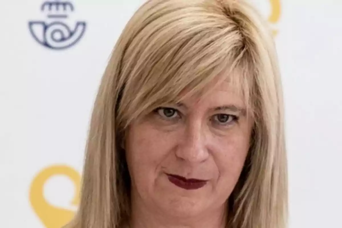 Mujer rubia de cabello lacio y labios pintados de rojo frente a un fondo blanco con logotipos amarillos y azules desenfocados Mujer rubia de cabello lacio y labios pintados de rojo frente a un fondo blanco con logotipos amarillos y azules desenfocados