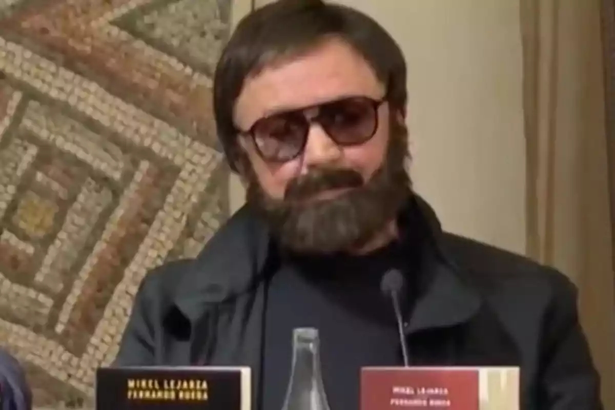 Un hombre con barba y gafas de sol está sentado frente a un micrófono con un libro en la mesa.