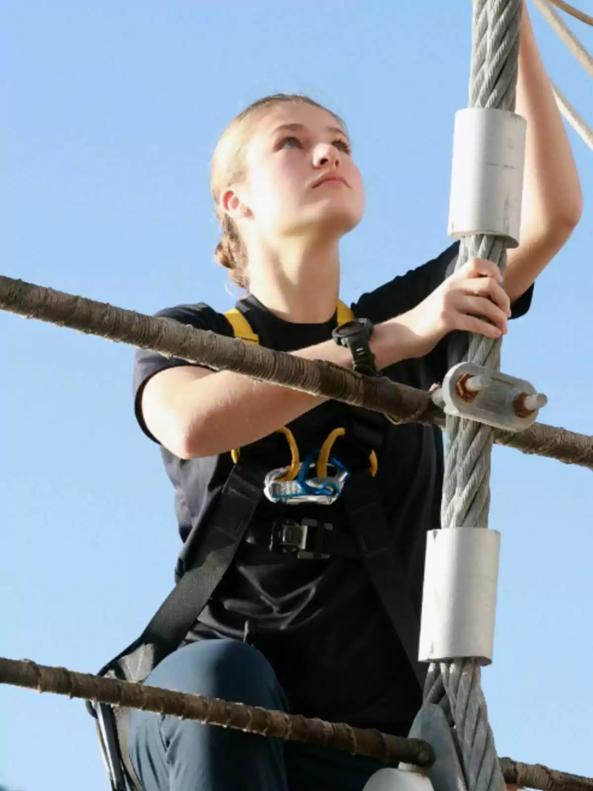Leonor con arnés de seguridad subiendo por una estructura de cuerdas bajo un cielo despejado. Leonor con arnés de seguridad subiendo por una estructura de cuerdas bajo un cielo despejado.