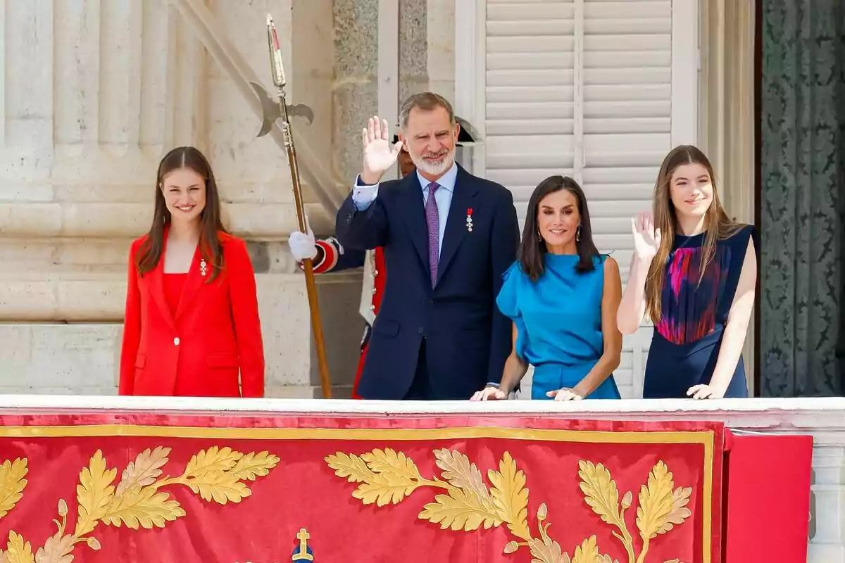 Leonor sonriendo en traje rojo junto a Felipe VI saludando en traje azul, Letizia en vestido azul y la infanta Sofía saludando en un vestido de colores en un balcón
