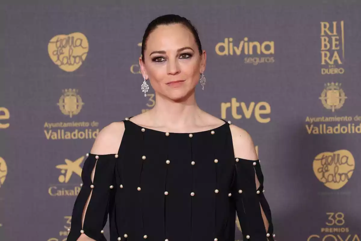 Una persona con un vestido negro decorado con perlas y pendientes brillantes posa frente a un fondo con logotipos de patrocinadores en un evento.