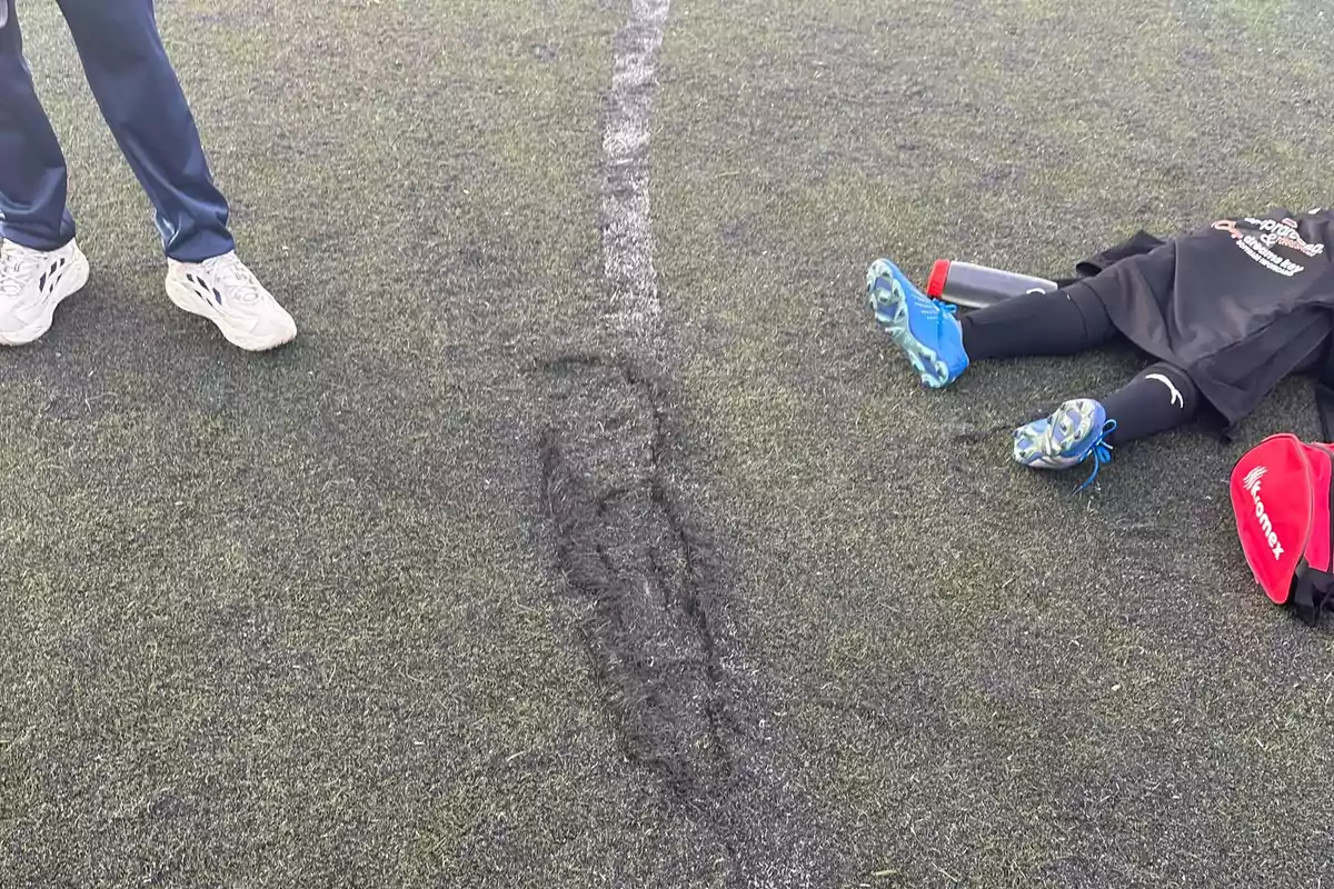 Una persona con zapatillas blancas de pie en un campo de césped artificial junto a otra persona acostada con zapatillas azules y una mochila roja.