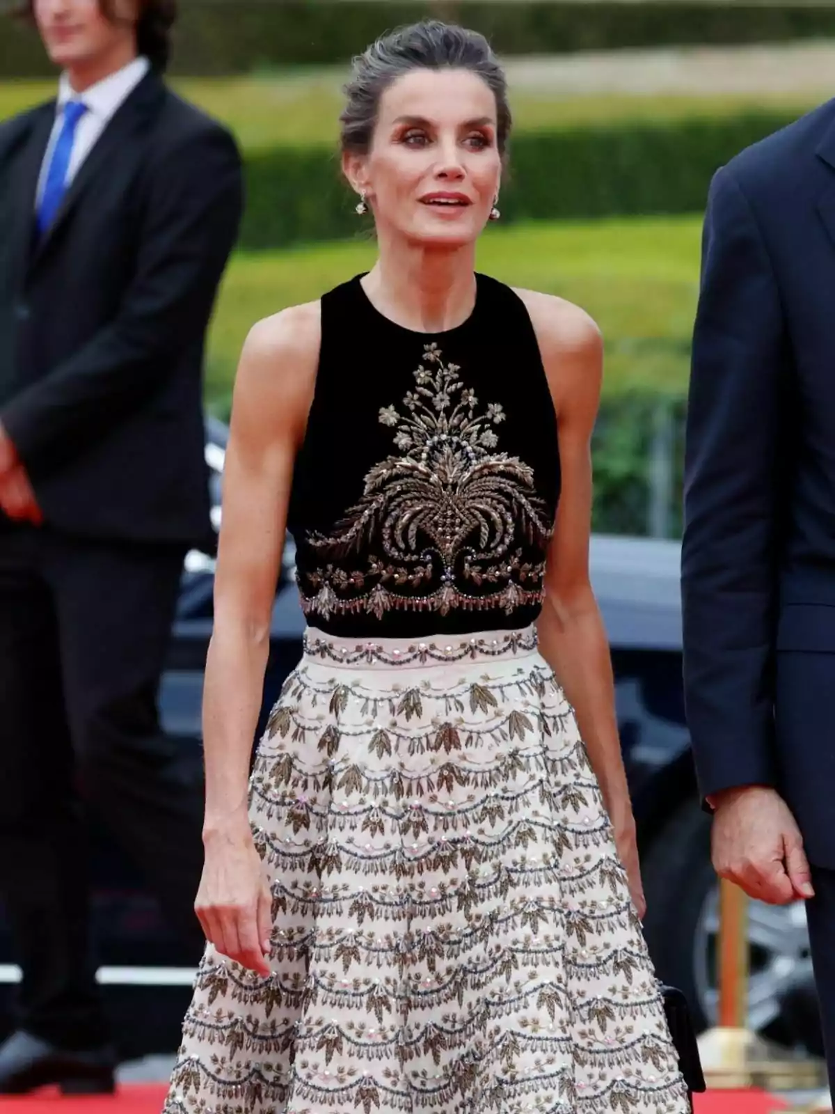 Letizia con un vestido elegante de dos piezas, con un top negro con detalles dorados y una falda blanca con patrones dorados, está de pie en un evento formal.