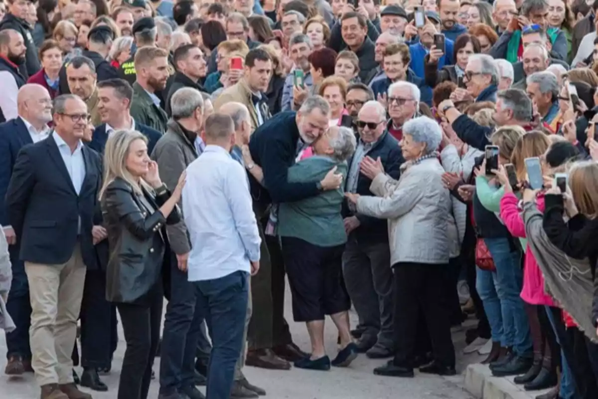 Un hombre abraza a una mujer mayor en medio de una multitud que observa y toma fotos.