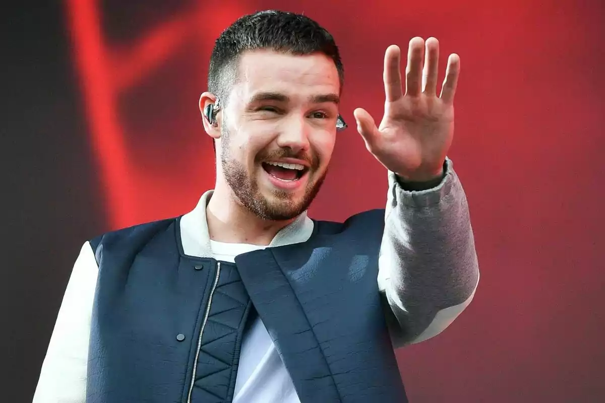 Liam Payne sonriente con barba y cabello corto saluda levantando la mano, usando una chaqueta azul y blanca, con un fondo rojo.