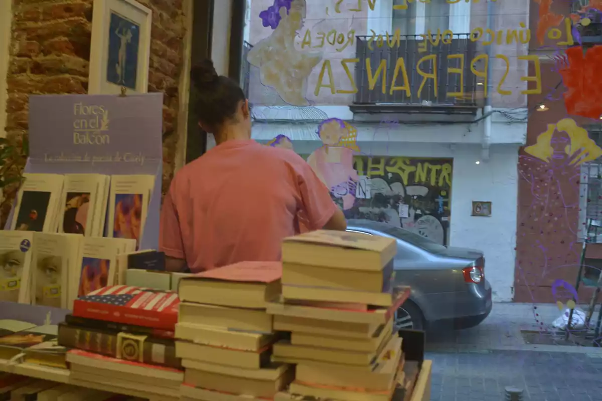 Una persona de espaldas con camiseta rosa está en una librería junto a una pila de libros y un expositor de poesía, mientras que en la ventana se ven dibujos y letras amarillas que dan al exterior de la calle.