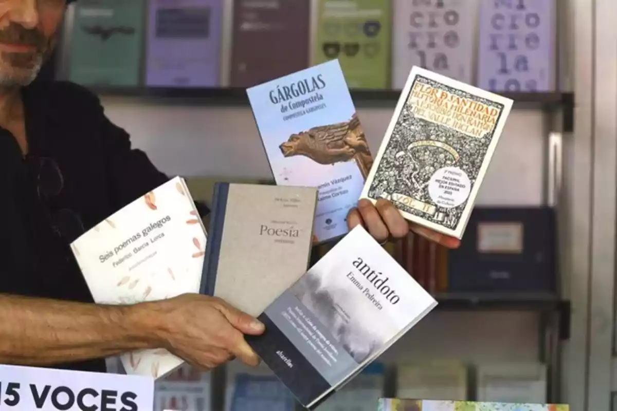 Persona sosteniendo cinco libros diferentes frente a una estantería llena de más libros en una feria o librería Persona sosteniendo cinco libros diferentes frente a una estantería llena de más libros en una feria o librería