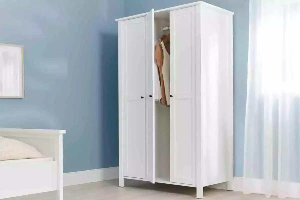 Armario blanco de dos puertas parcialmente abierto con ropa colgada en su interior en una habitación de paredes azules y cama blanca Armario blanco de dos puertas parcialmente abierto con ropa colgada en su interior en una habitación de paredes azules y cama blanca