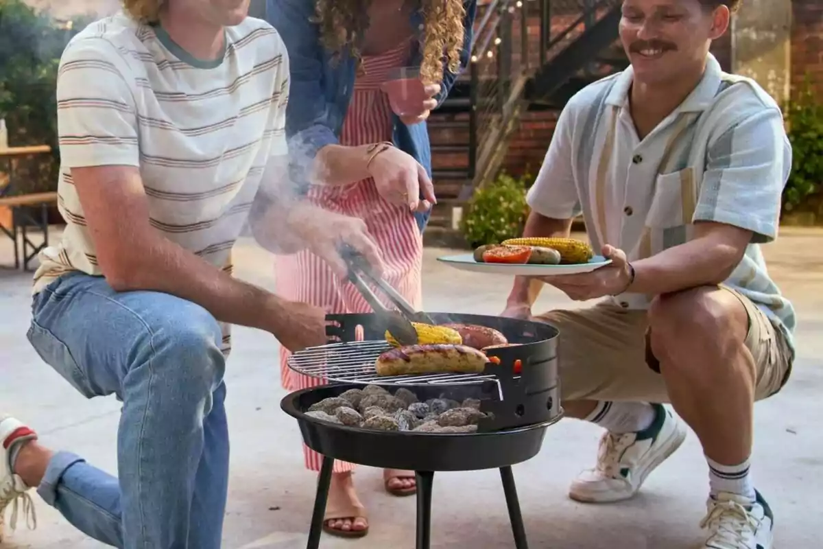 Tres personas disfrutan de una parrillada al aire libre cocinando salchichas y elotes en una parrilla portátil Tres personas disfrutan de una parrillada al aire libre cocinando salchichas y elotes en una parrilla portátil