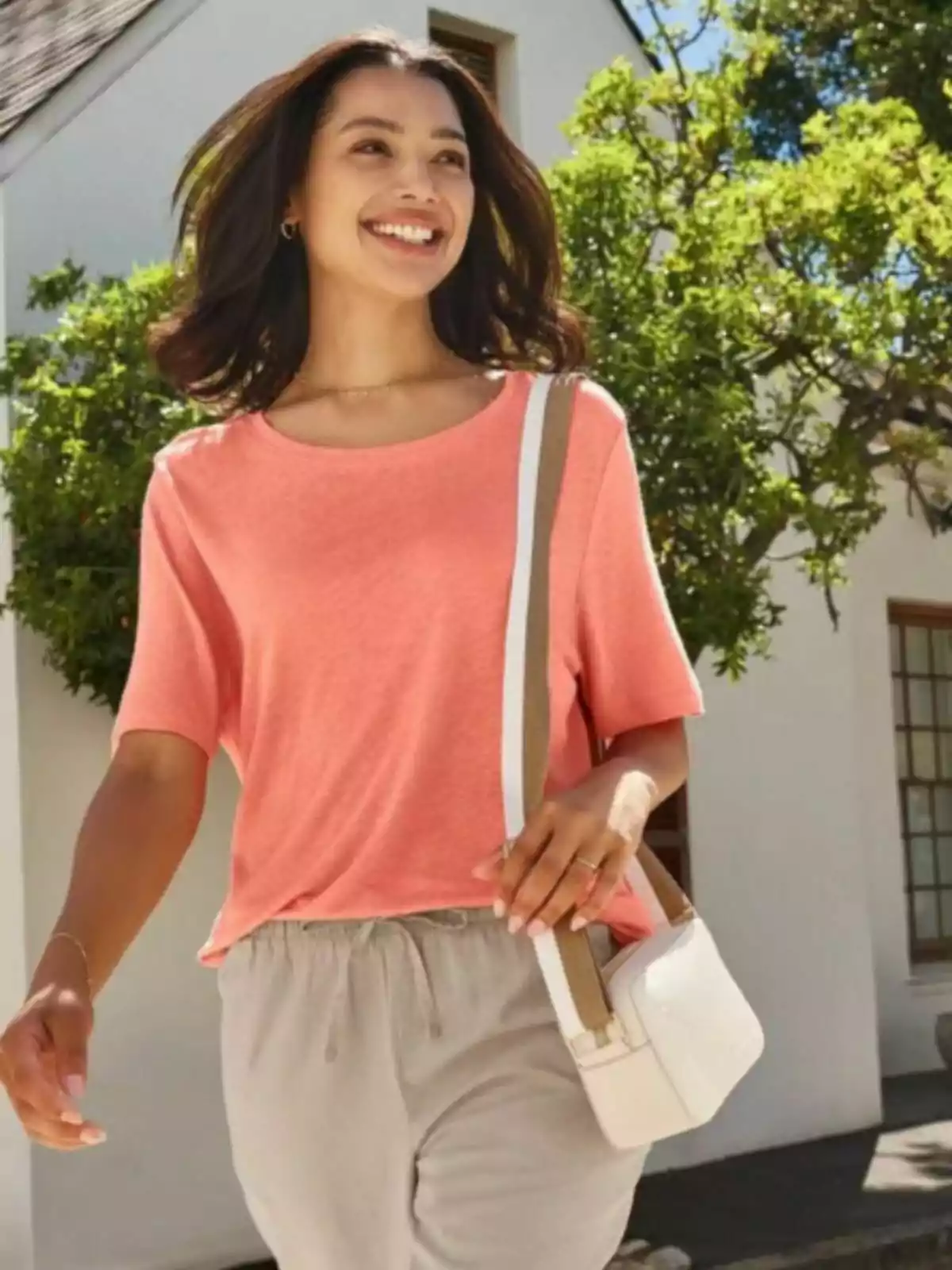 Una persona sonriente camina al aire libre con una camiseta color salmón y un bolso blanco, rodeada de vegetación y una casa blanca de fondo.