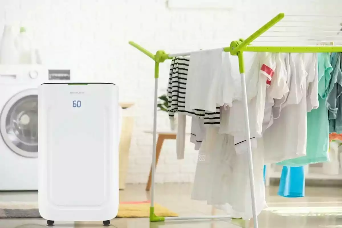 Un deshumidificador blanco junto a un tendedero con ropa secándose en una habitación con lavadora al fondo Un deshumidificador blanco junto a un tendedero con ropa secándose en una habitación con lavadora al fondo